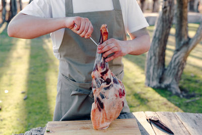 Anonymous male cook in apron standing at table in countryside with raw leg of lamb and preparing meat for hanging on metal hook