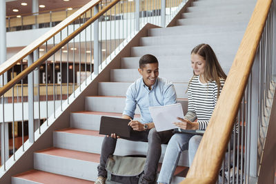 Smiling friends discussing over documents while sitting on steps in university