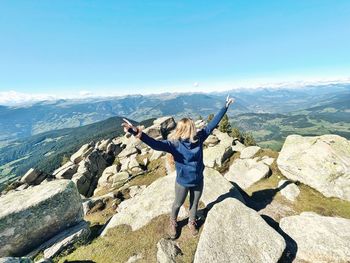 View of young woman standing happily on mountain against clear blue sky.