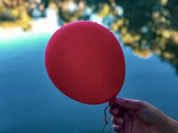 Close-up of hand holding red balloon against reflections in lake water.