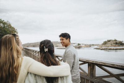 Smiling young man with female friends walking on bridge near lake