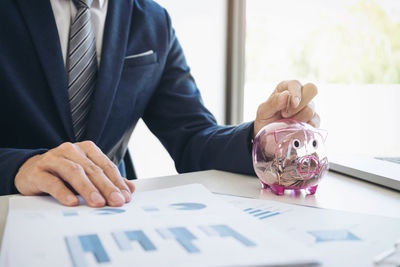 Midsection of businessman putting coin in piggy bank while sitting at table in office