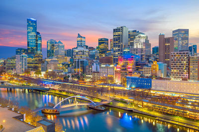 Illuminated bridge over river by buildings against sky at night