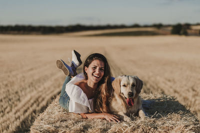 Happy mid adult woman with dog lying on hay at agricultural field