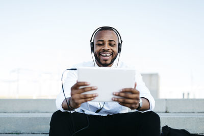 Happy man sitting on stairs wearing headphones looking at tablet