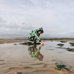 Boy crouching at beach against sky