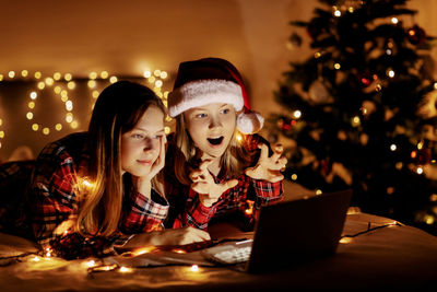 Two charming sisters lie on the bed with a laptop on christmas or new year's eve 