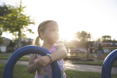Cute girl looking away while holding curved blue metal at playground during sunset