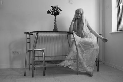 Young woman wearing dress sitting on chair by table against wall at home