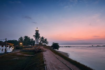 Illuminated building by sea against sky during sunset