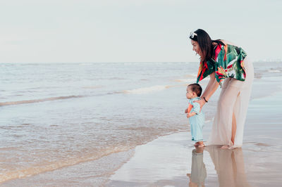 Mother with baby boy standing at beach against clear sky