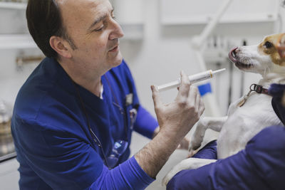 Side view of crop doctors in uniform holding dog and pouring medicament from syringe in mouth in medical room