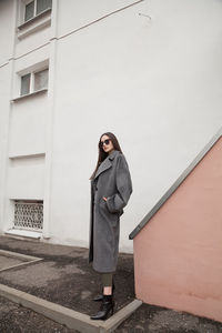 Full length side view portrait of young woman in grey coat and suit against wall outdoors, outwear 