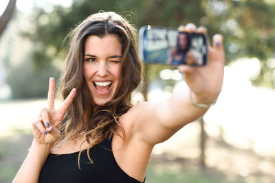 Cheerful young woman gesturing peace sign while taking selfie on smart phone