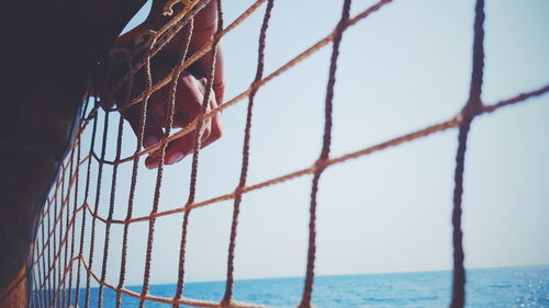 Cropped image of hands on net by sea against clear sky