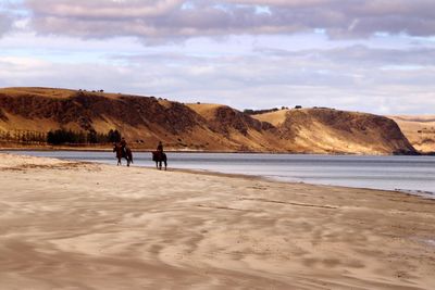 Men riding horses at beach against cloudy sky