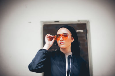 Portrait of young woman wearing sunglasses standing against wall