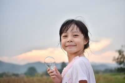 Portrait of smiling girl holding light bulb against cloudy sky