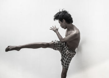 Shirtless young man kicking while standing against white background