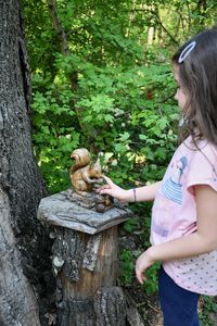 Side view of girl playing with squirrel statue by tree trunk