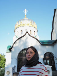 A girl of european appearance is standing in a kerchief at a white orthodox church. 