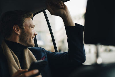 Smiling businessman with phone looking through window while sitting in taxi