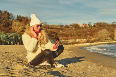 A woman in a fur coat and hat sits on a sandy beach and listens to music on a smartphone.