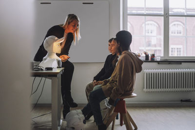 Smiling teacher discussing with multiracial students sitting in front of illuminated social robot in innovation lab