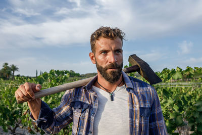 Young bearded hispanic male farmer in casual clothes looking away while standing amidst rows of plants with hoe in hand during work in countryside