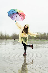 Beautiful brunette woman in yellow raincoat holding rainbow umbrella dancing out in the rain