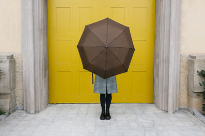 Woman holding umbrella while standing against yellow door