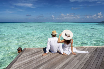 Rear view of couple sitting on pier looking at sea against sky