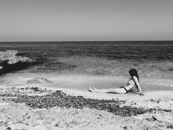 Side view of young woman sitting on shore at beach against sky