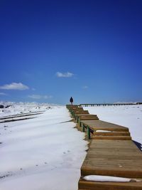 Person walking on pier over snowy landscape against blue sky