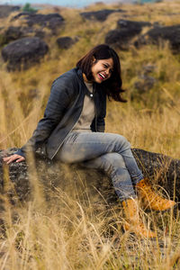 Full length of cheerful young woman sitting on rock at farm