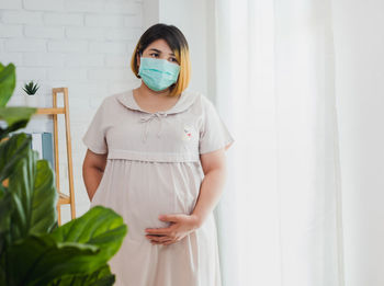 Pregnant woman wearing mask looking away while standing against wall