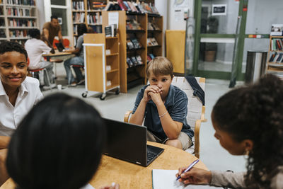 Thoughtful teenage boy sitting with laptop near table in library at junior high school