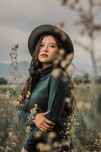 Young woman wearing hat looking away while standing on field against cloudy sky