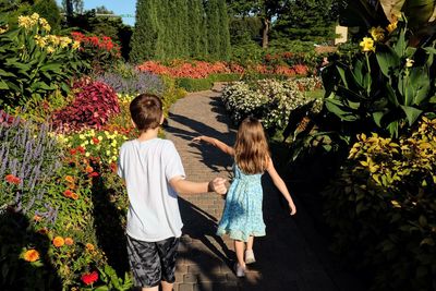 Rear view of siblings walking on footpath amidst flowering plants at garden