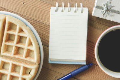 Directly above shot of spiral notebook with black coffee and waffle by gift on table