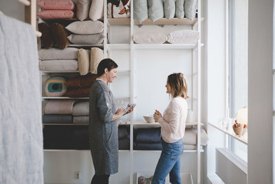 Side view of female design professionals discussing against shelves in upholstery workshop