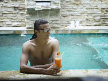 Shirtless young man holding drink while swimming in pool