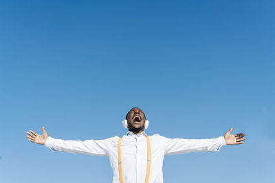 Carefree man screaming with arms outstretched in front of blue sky