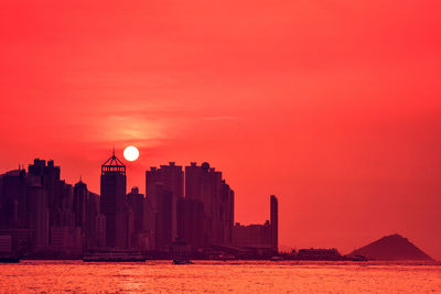 Silhouette buildings by sea against orange sky