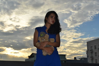 Low angle view of thoughtful woman holding teddy bear against cloudy sky