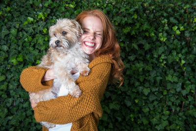 Cheerful woman holding dog while standing by ivies