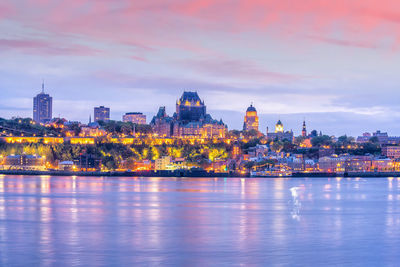 Illuminated buildings by river against cloudy sky