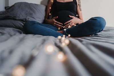 Midsection of pregnant young woman sitting with string lights on bed at home