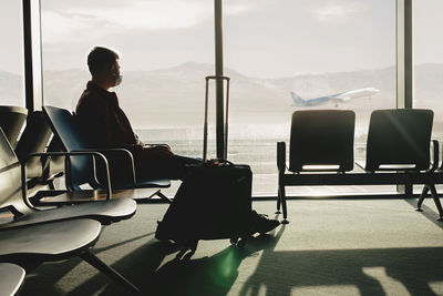 Man wearing a mask waiting his flight at the gate in the airport. looking to the taking off plane. 