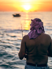 Rear view of shirtless man fishing in sea against sky during sunset